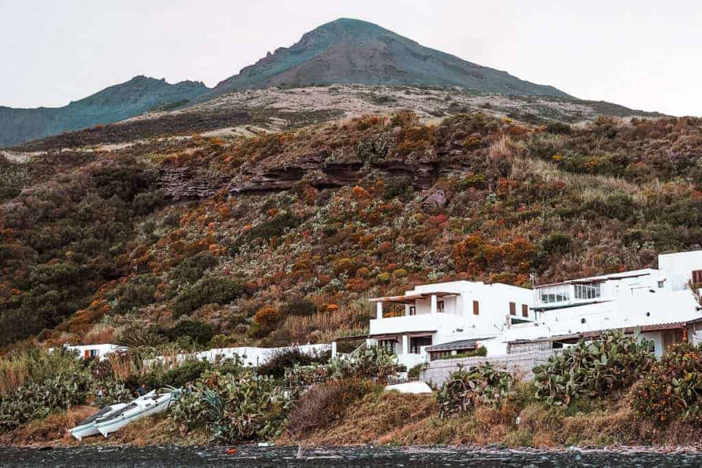 A collection of the best landmarks in Italy and one is Stromboli. And in this picture you see an active volcano in the background that is covered in grass and that has an expansive white building at it's base.