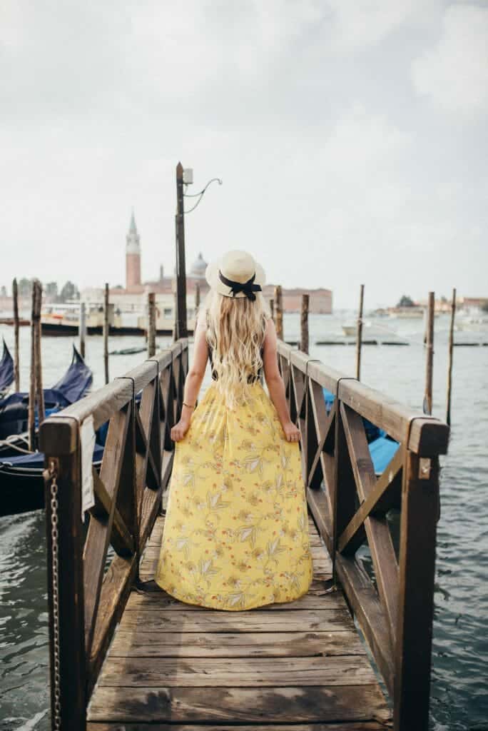 A woman stands with long blonde hair and a hat with a black bow in the back . She has her hands holding a long yellow skirt as she stands on a dock in Venice that is in the water and surrounded by gondolas that are perfect to ride in if you're wondering how to spend 2 days in Venice.