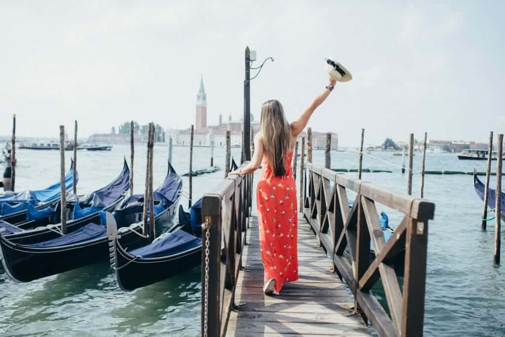 Paula standing in a red dress with a hat raised in her right hand as she stands on a dock and looks out on Venice during her Venice Itinerary. Gondolas sit next to her docced on her left.
