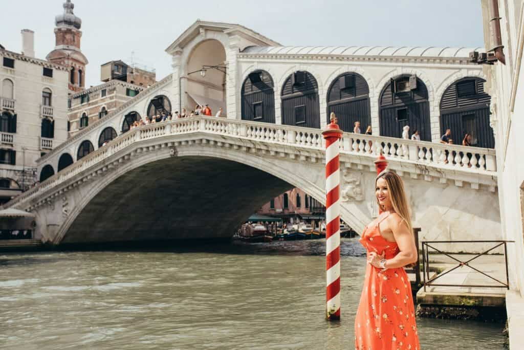 Paula stands by the canal in front of Rialto Bridge during her 2 Day Itinerary in Venice. She is wearing a red dress and there is a red and white striped pole in the water to her left.