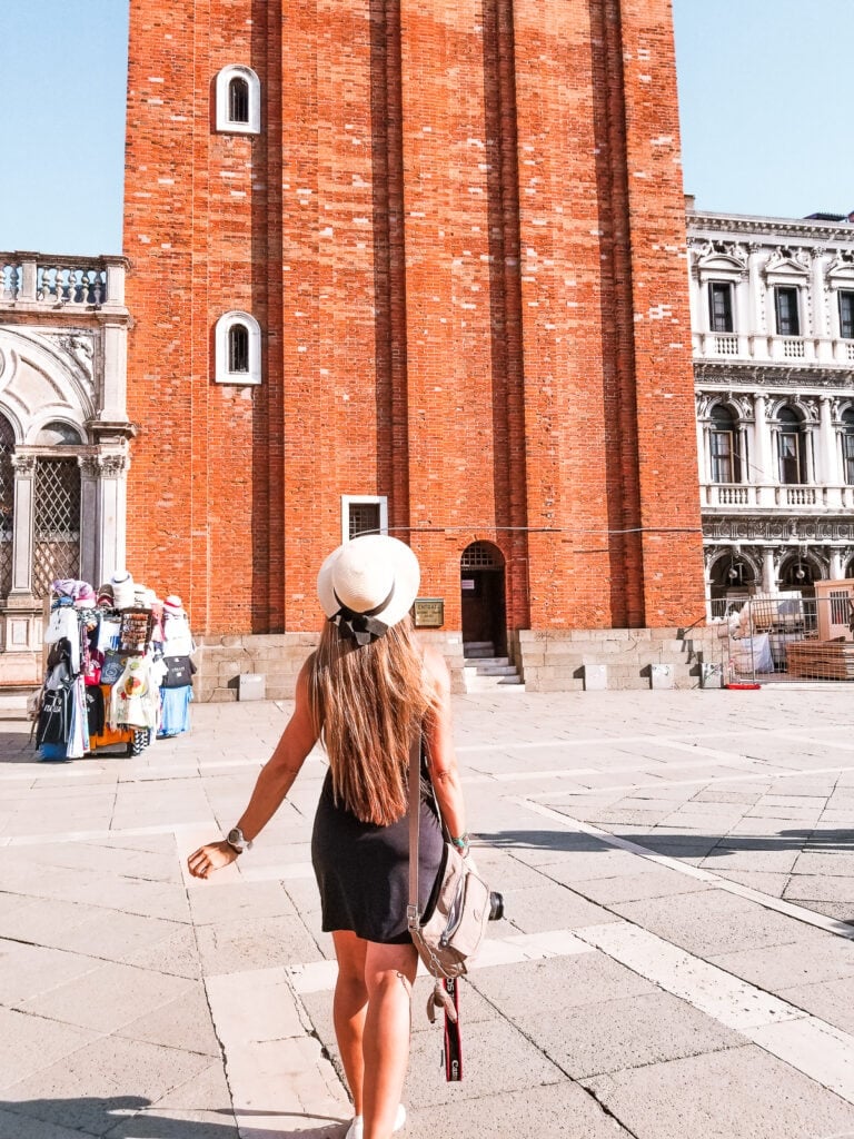 Paula stands in front of the base of Saint Marks’ Campanile. She has a wide brim het with a bow on and is in a dress with her back to the camera.