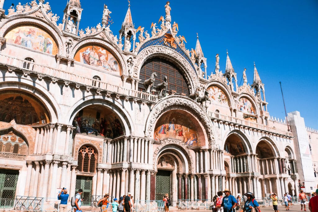 Elaborate exterior of St Mark’s Basilica with frescoes over the doors and lots of arched windows and archways.